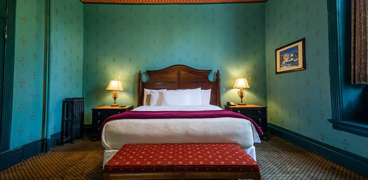 A wide view of a guest room with a large bed, two nightstands, a bench at the foot of the bed, and teal wallpaper.