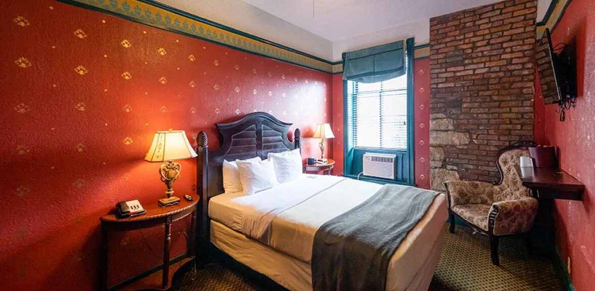 A wide view of a hotel room with a bed, nightstand, lamp, chair, and window