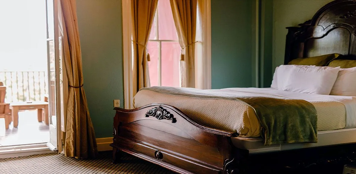 A close-up view of a bed in a room with a wooden frame, white bedding, and a green bedspread, with a window and balcony in the background.