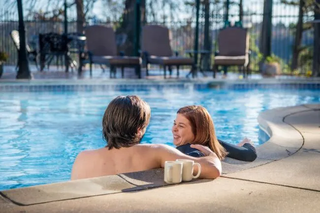 A couple relaxes in a pool with coffee mugs, surrounded by lounge chairs and trees.