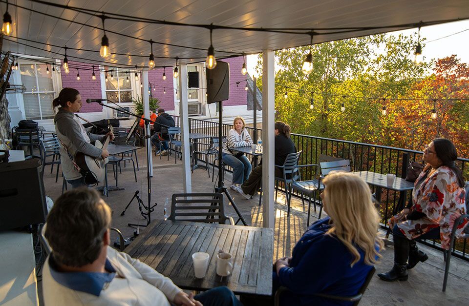A group of people sitting at tables on a covered patio with string lights, listening to a woman playing guitar.