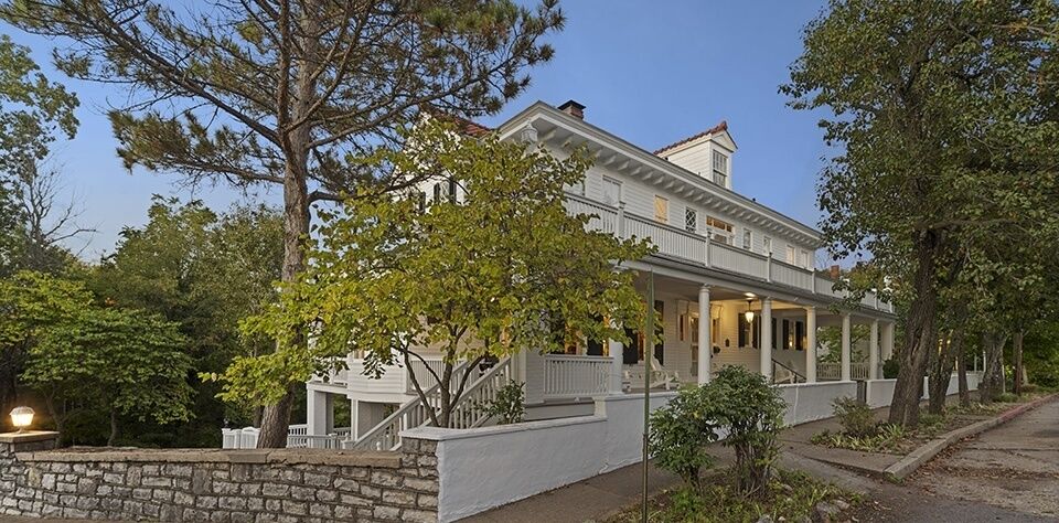 A large white hotel building with a porch and balcony surrounded by trees